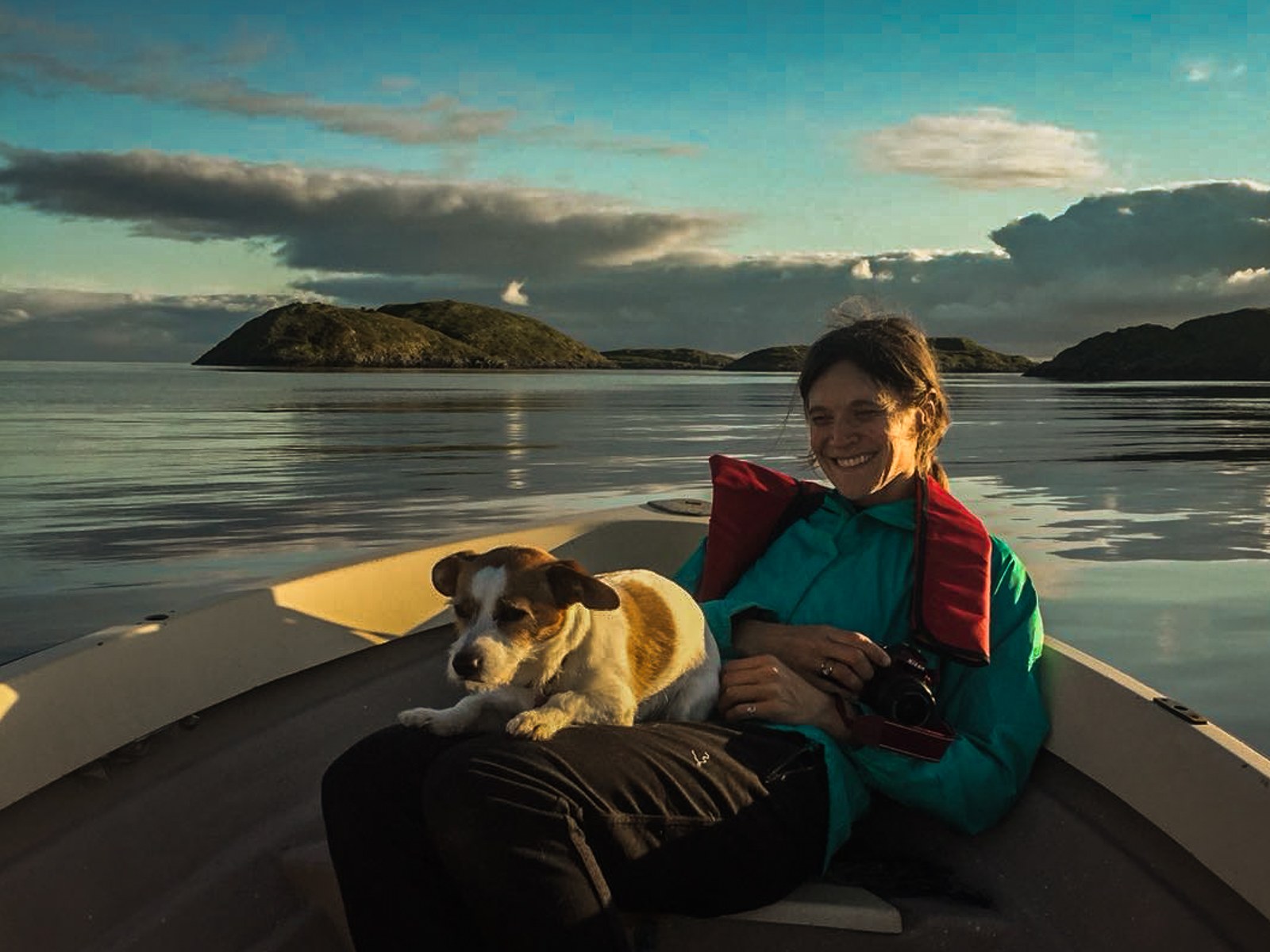 Andrea smiling in a boat with her dog Leo and a camera, golden evening light on the water.