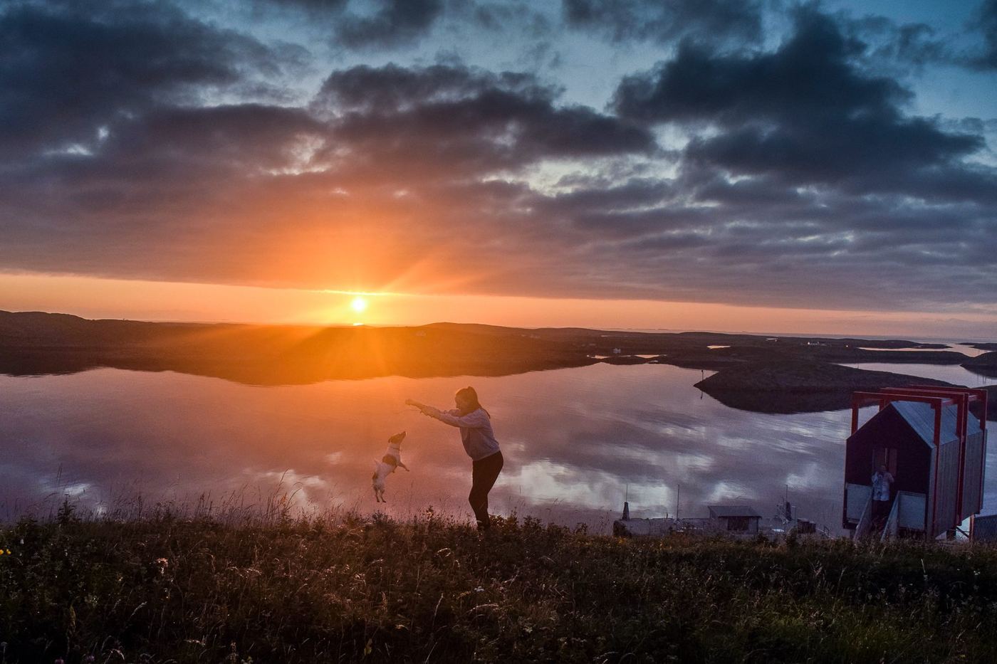 Andrea jugando con su perro Leo en una colina sobre el fiordo de Fleinvær, Noruega, al atardecer, con una pequeña cabaña roja cerca.
