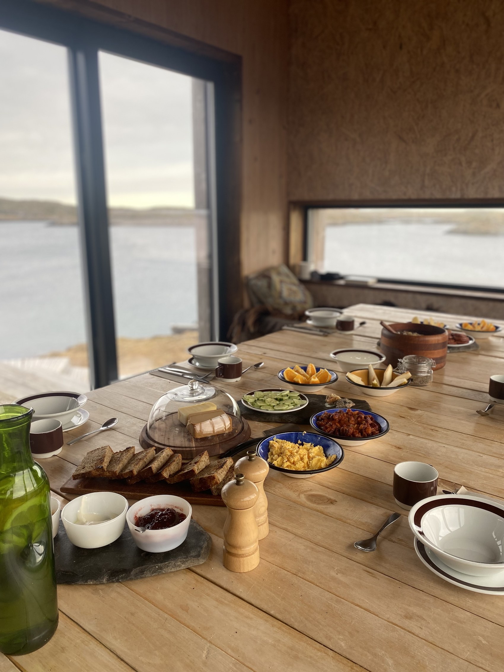 A long breakfast table set with bread, jam, fruit and coffee, looking out over the water at Fleinvær.
