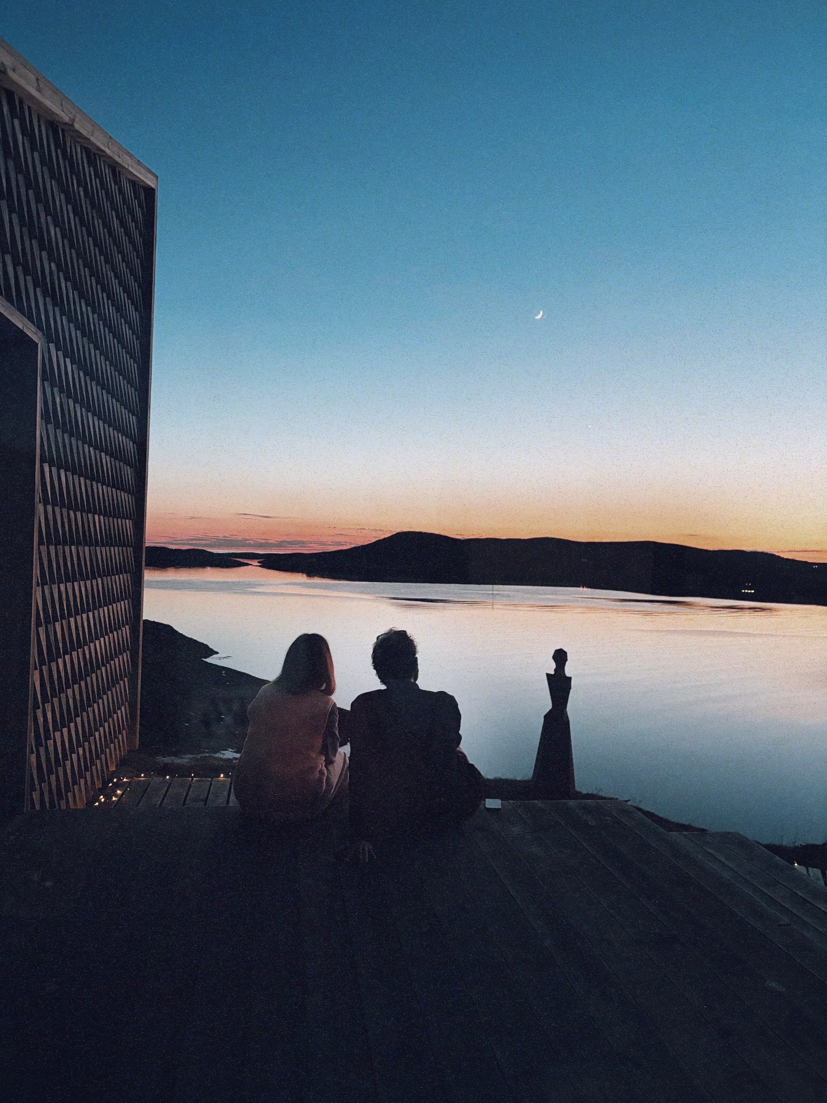 Two figures sitting at dusk on a wooden deck, watching the fjord with a small sculpture beside them and a crescent moon overhead.