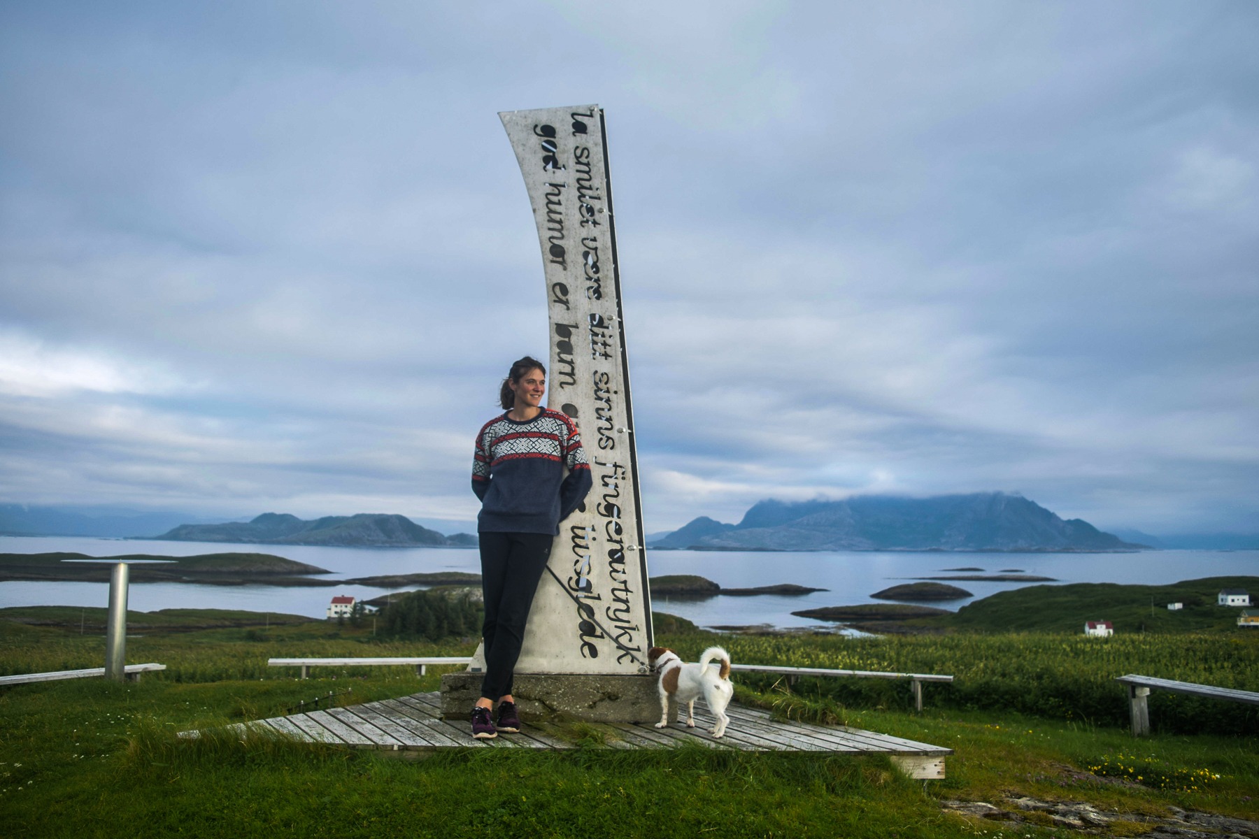 Andrea junto a una alta escultura de vela de madera con vistas al fiordo y las islas de Fleinvær, Noruega, con su perro Leo cerca.
