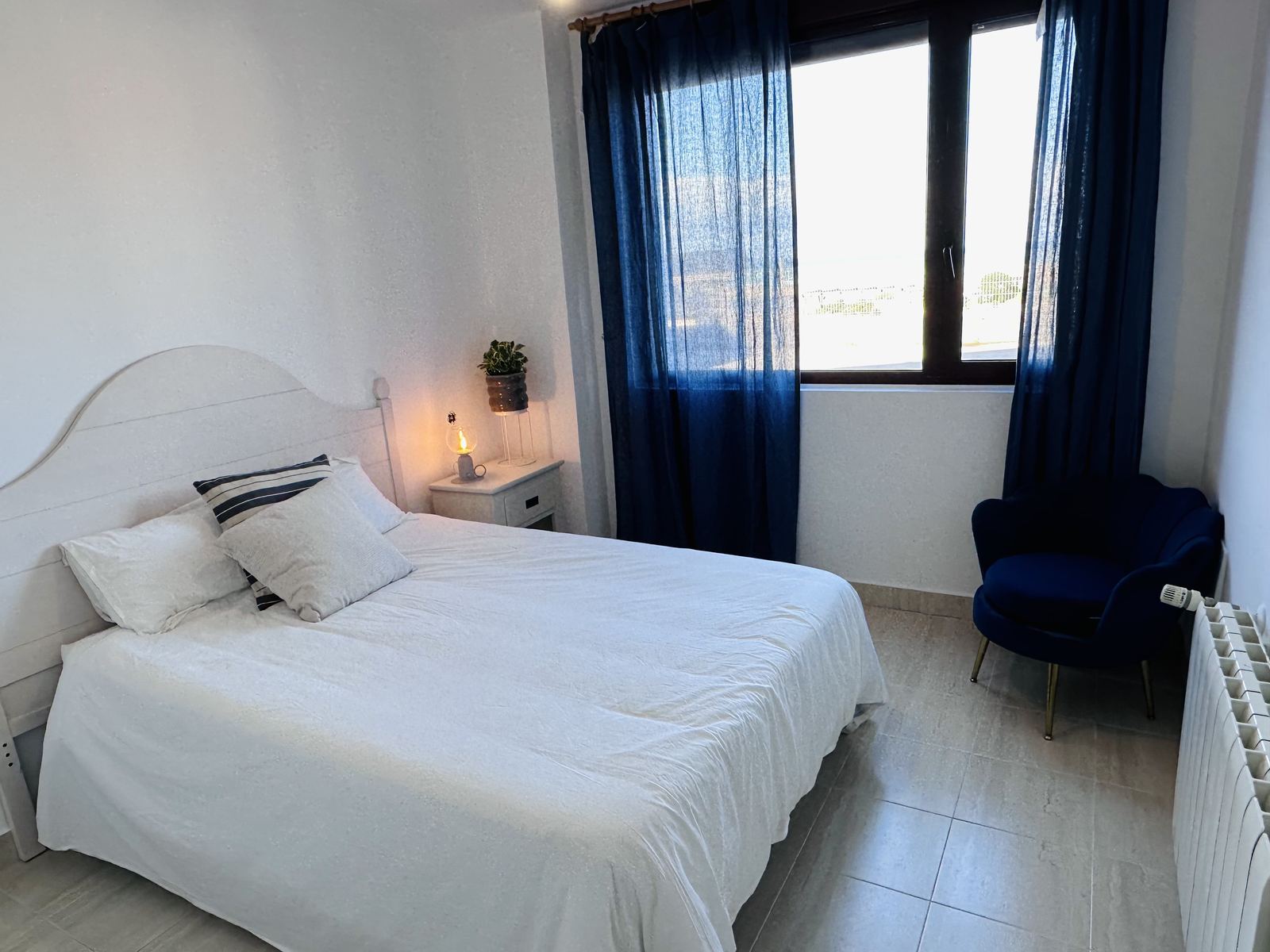 A bedroom in the apartment, with white bedding, a white headboard and navy linen curtains.