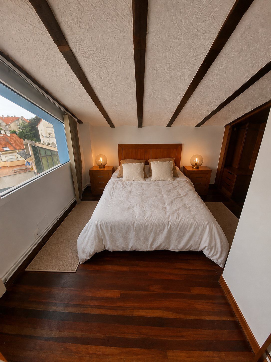 An attic bedroom with wooden beams, a wardrobe and a rooftop view through the window.