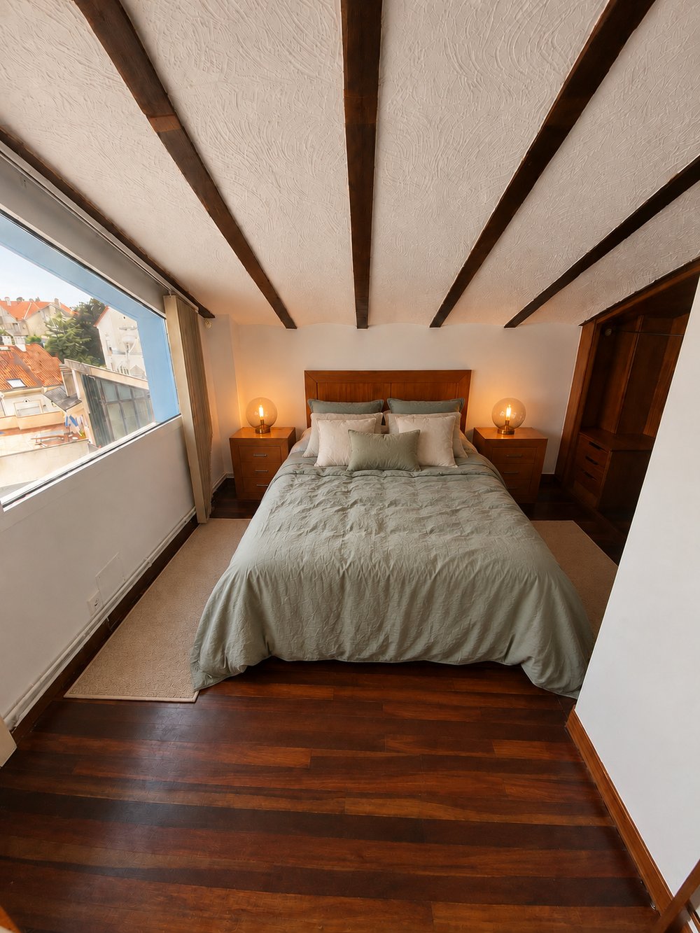 A loft bedroom with beamed ceilings, sage green bedding, and a window looking out over the rooftops.