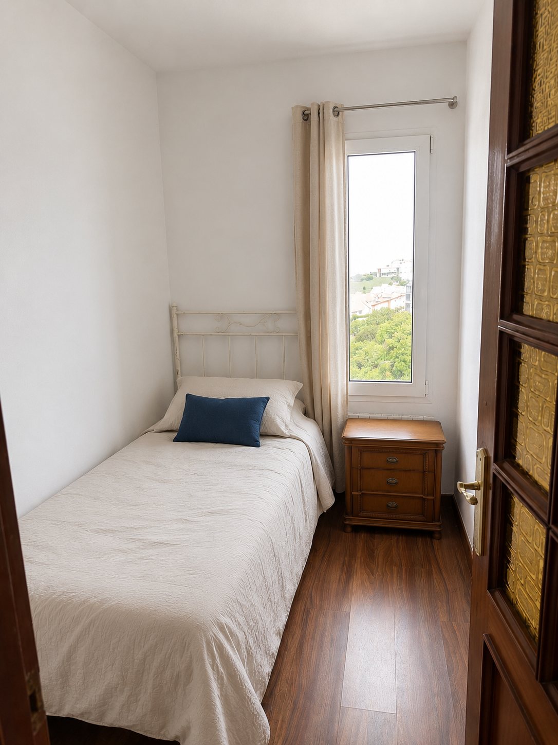 A narrow single bedroom with a navy accent pillow and dark wood floor.