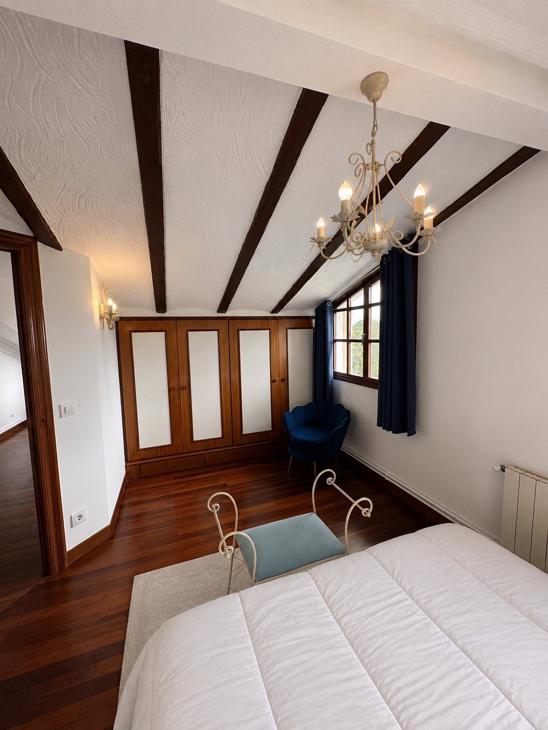 A cream chandelier above a made bed, with a built-in wardrobe and a blue velvet chair in the corner.