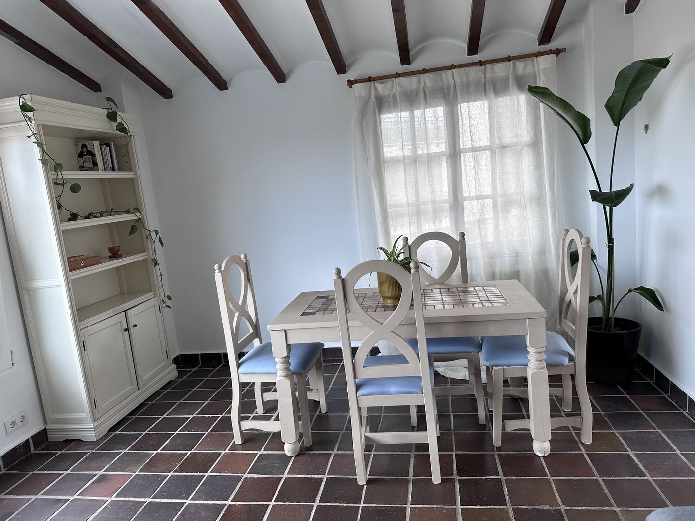 A dining table and chairs beneath beamed ceilings, with a tall plant and sheer curtains by the window.