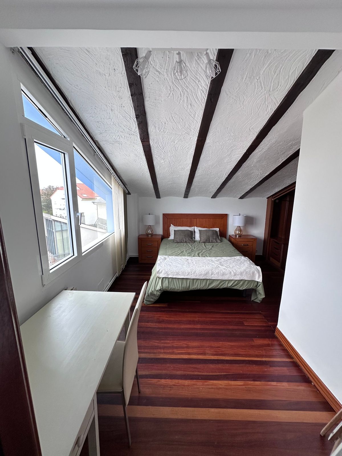 A bedroom under exposed beams, with a wooden headboard, green bedding, a desk by the window and dark wood floors.