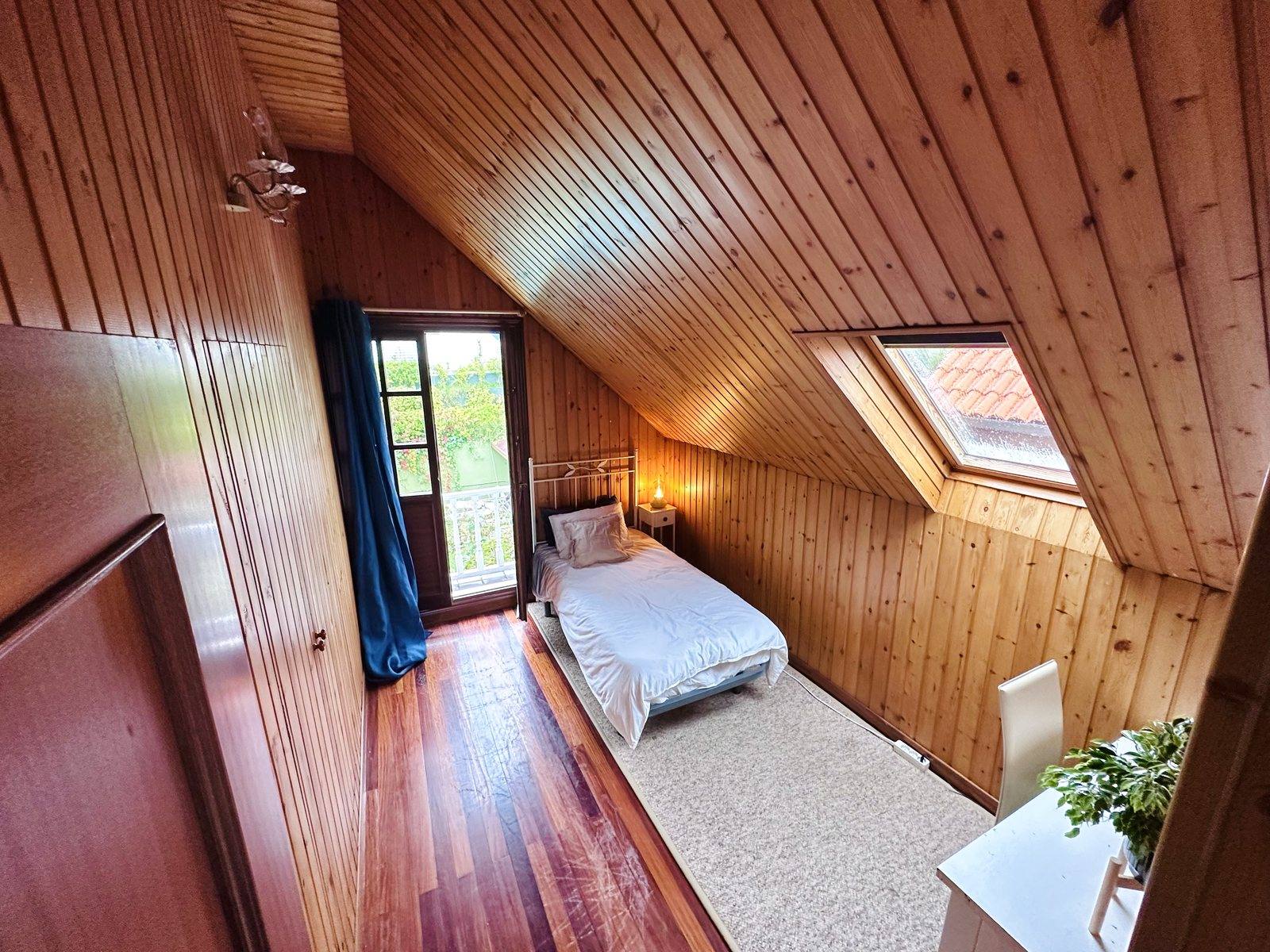 A wood-panelled attic bedroom seen from the doorway: a single bed, a balcony door with a navy curtain, a skylight and a desk.