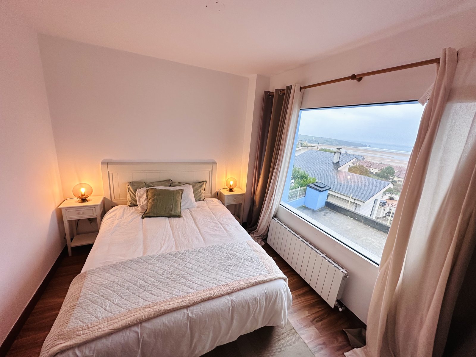 A bright bedroom with a wide picture window looking over the rooftops toward the sea.