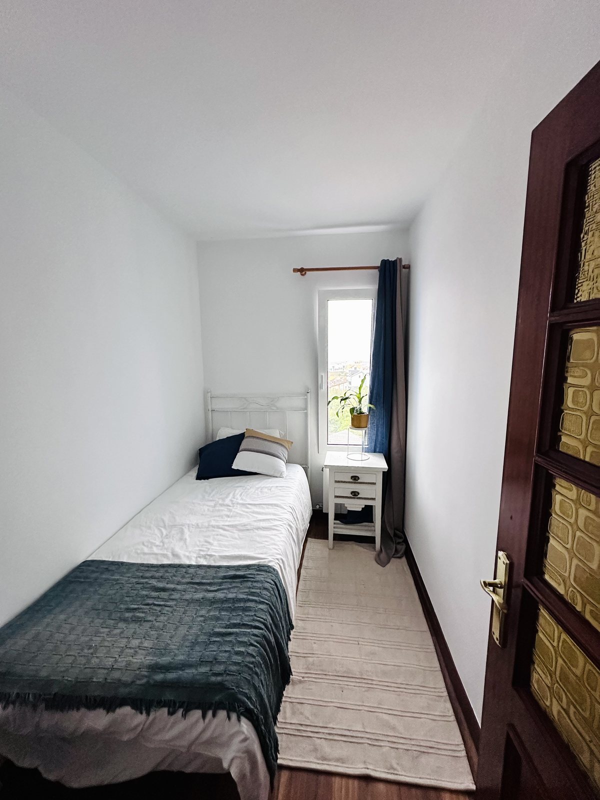 A cosy single bedroom with a white iron bedstead, a teal throw and a small window.