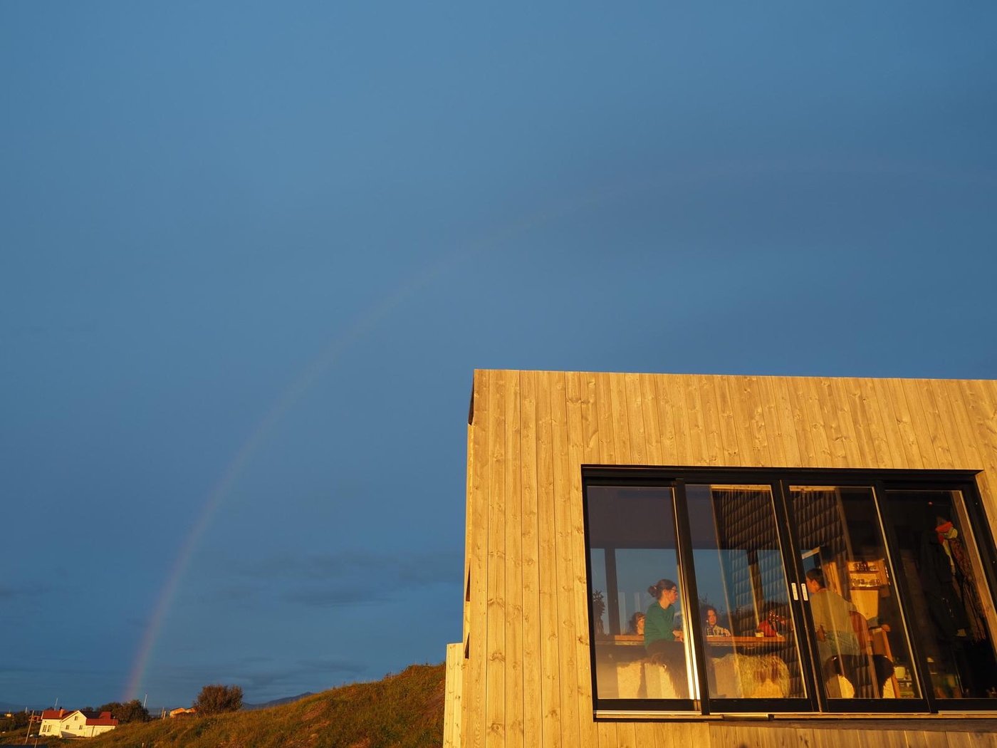 A wooden cabin at golden hour, with a rainbow overhead and guests gathered at the table inside.