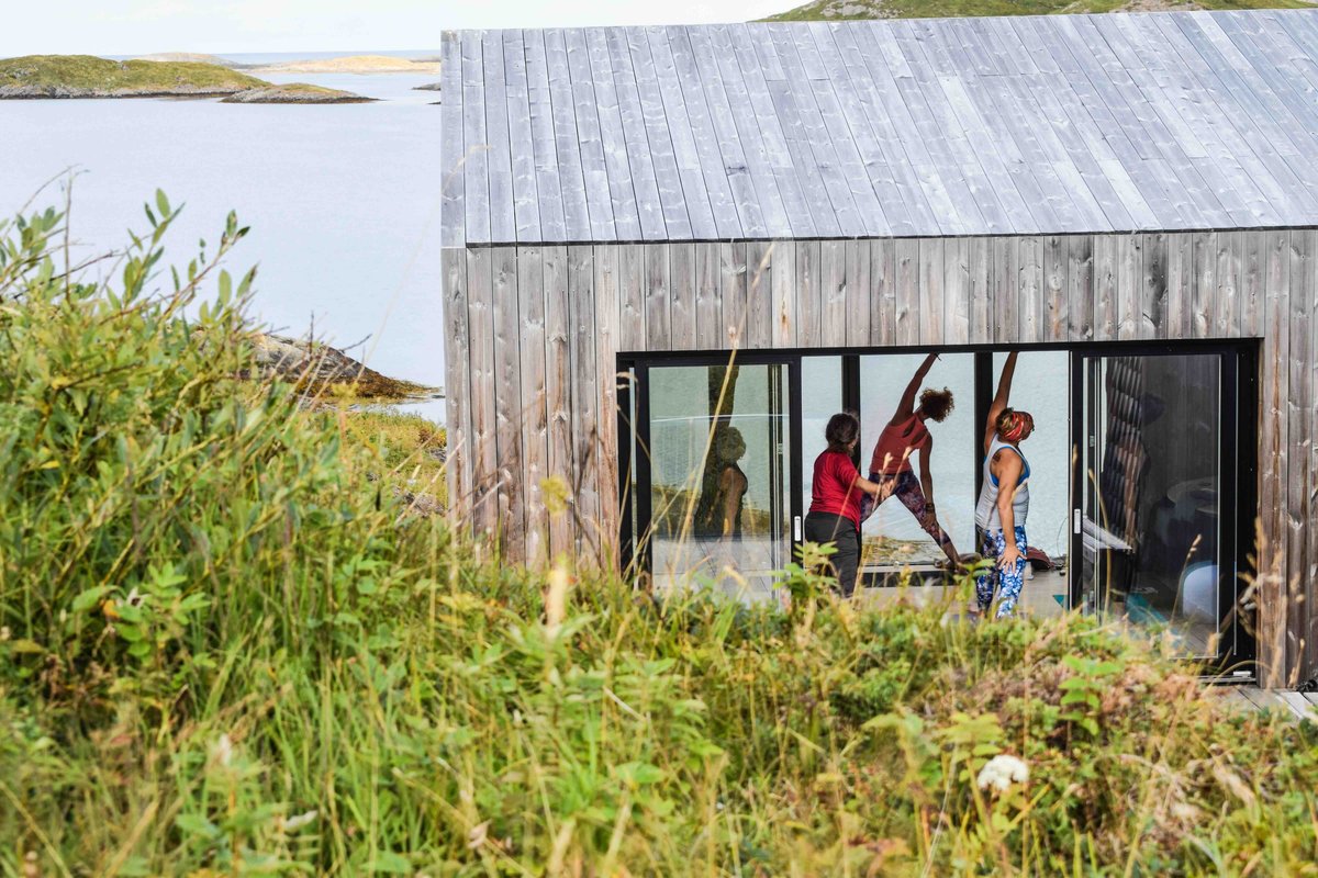 Three women stretching together on the deck of a cabin overlooking the water.