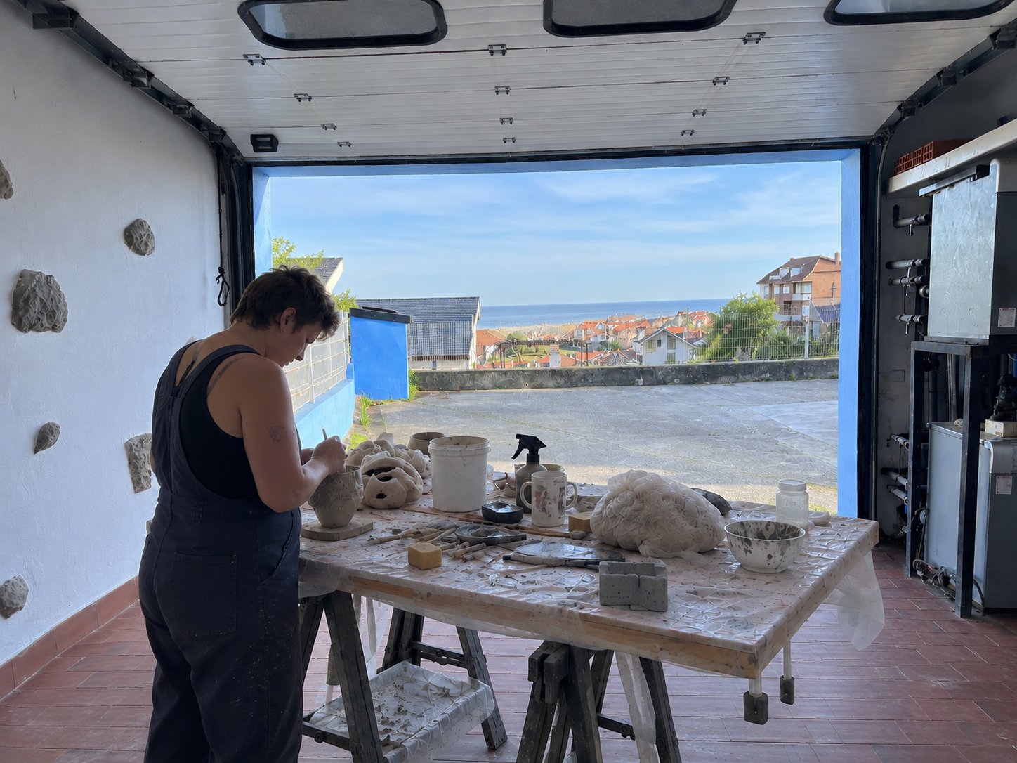 Una artista trabajando en una mesa cubierta de arcilla en un estudio abierto al mar, dando forma a una pieza de cerámica.