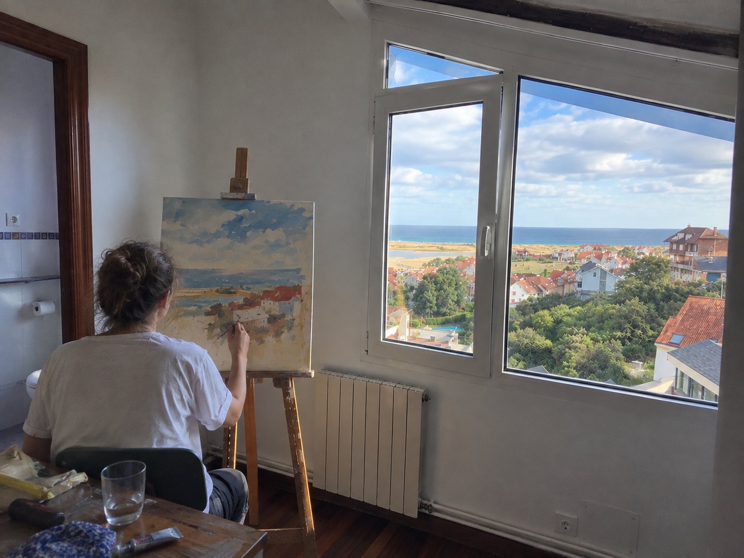 Una artista pintando en un caballete en una habitación luminosa con vistas a la costa, con material de pintura sobre una mesa cercana.