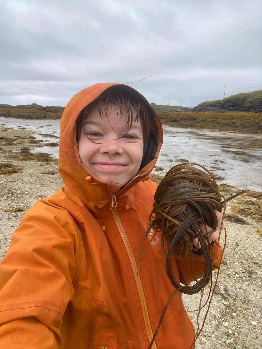 Una persona con un chubasquero naranja en la playa, sosteniendo un manojo de algas recogidas.